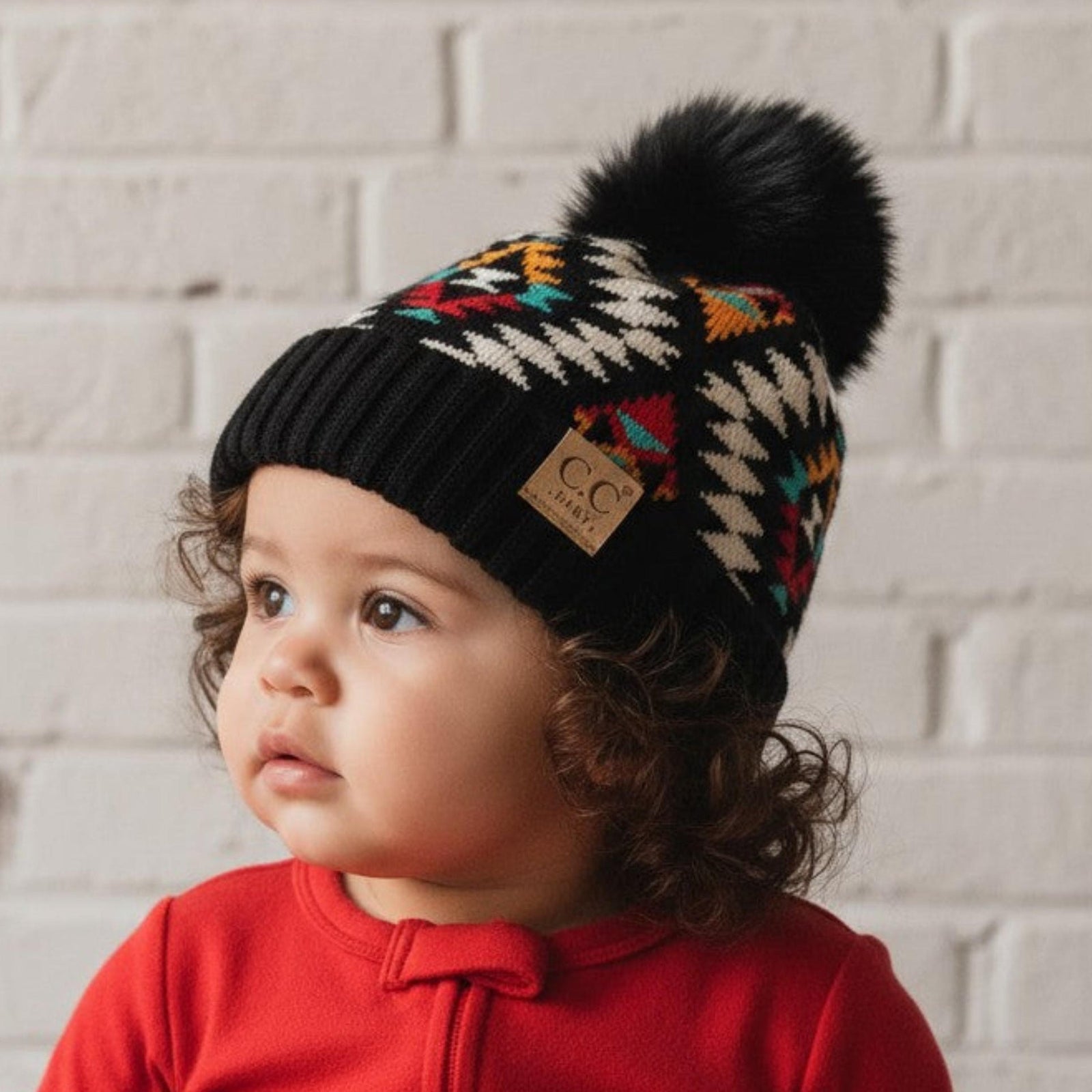 Child wearing a colorful knit hat with a pom-pom against a light brick wall.