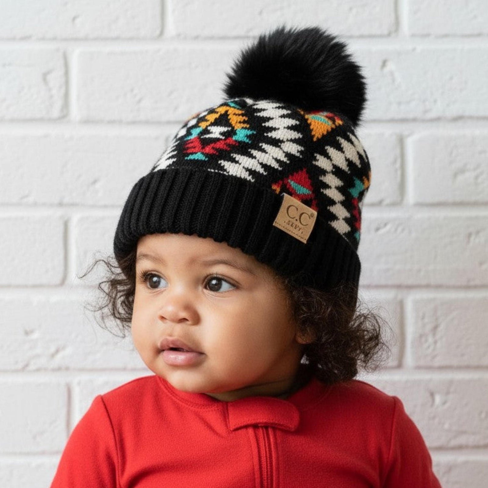 Child wearing a colorful knit hat with a pom-pom and a red outfit against a white brick wall.
