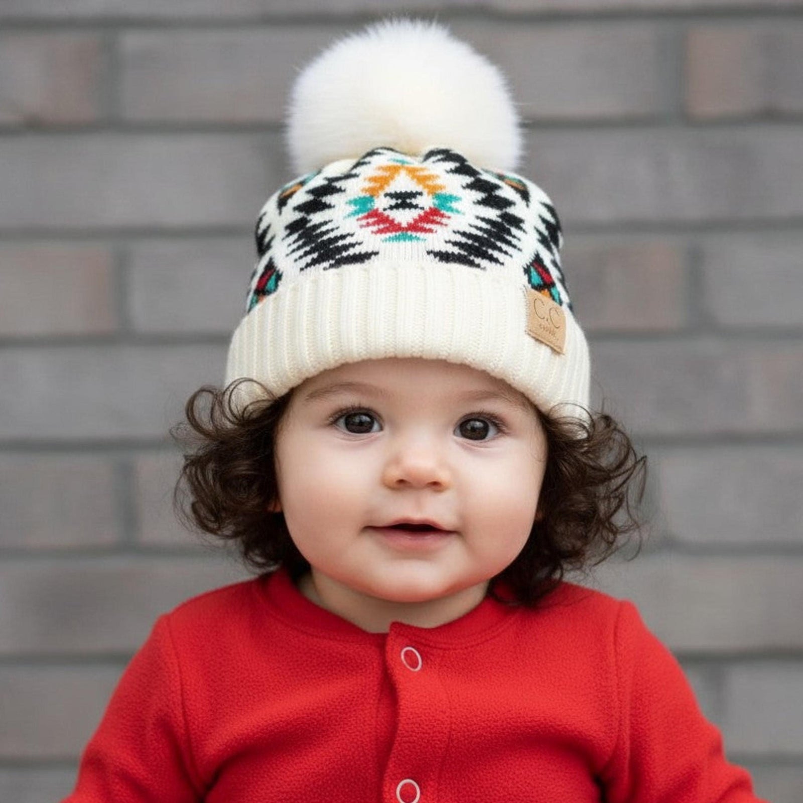 Child wearing a red outfit and patterned hat with a pom-pom against a brick wall.