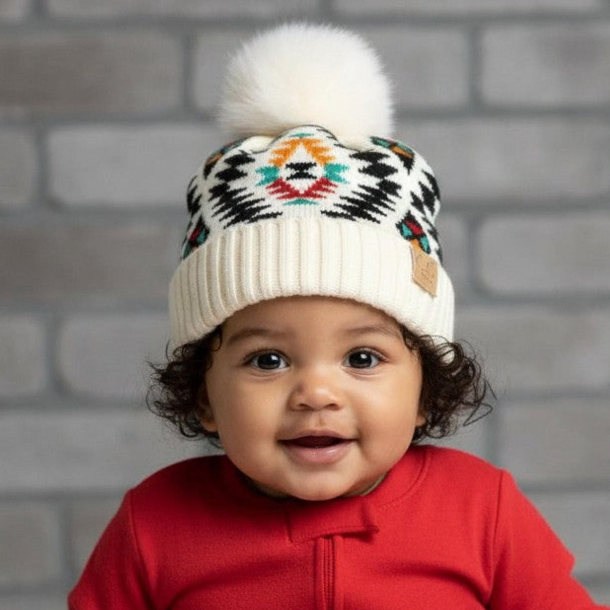 Baby wearing a red outfit and patterned hat sitting against a brick wall.