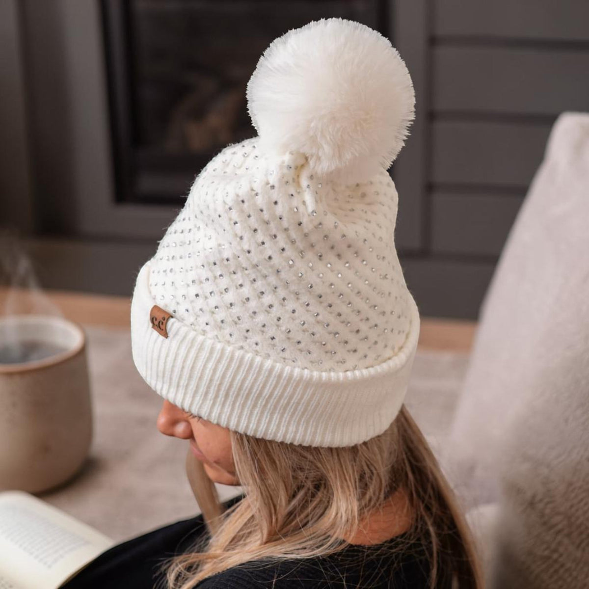 Truly Contagious white C.C beanie with allover rhinestones and a large faux fur pom pom on a woman reading