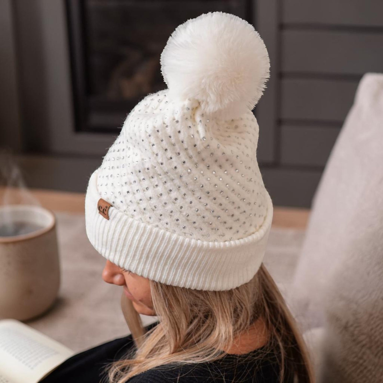 Truly Contagious white C.C beanie with allover rhinestones and a large faux fur pom pom on a woman reading