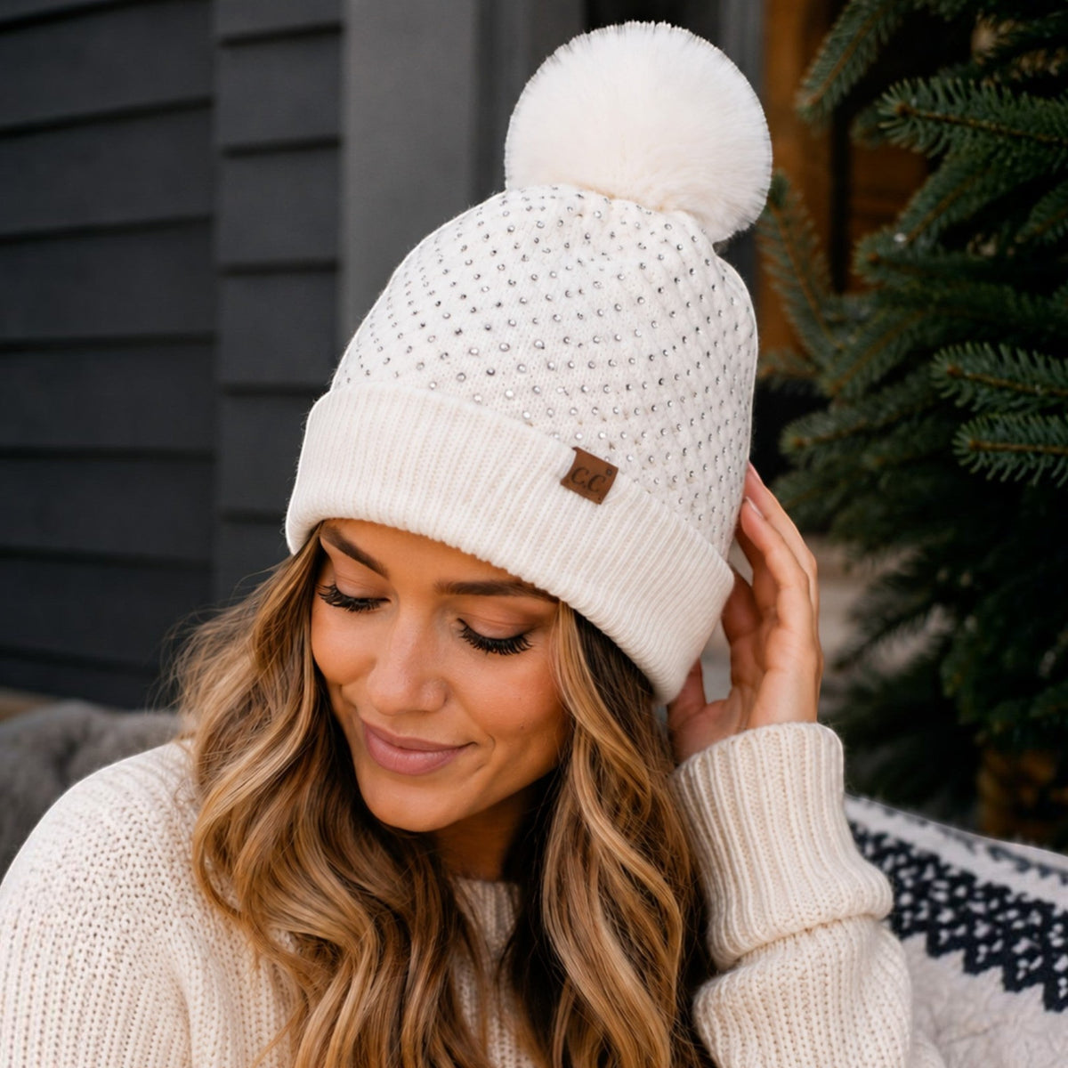 Woman wearing a white Truly Contagious C.C beanie with allover rhinestones and a large faux fur pom pom.